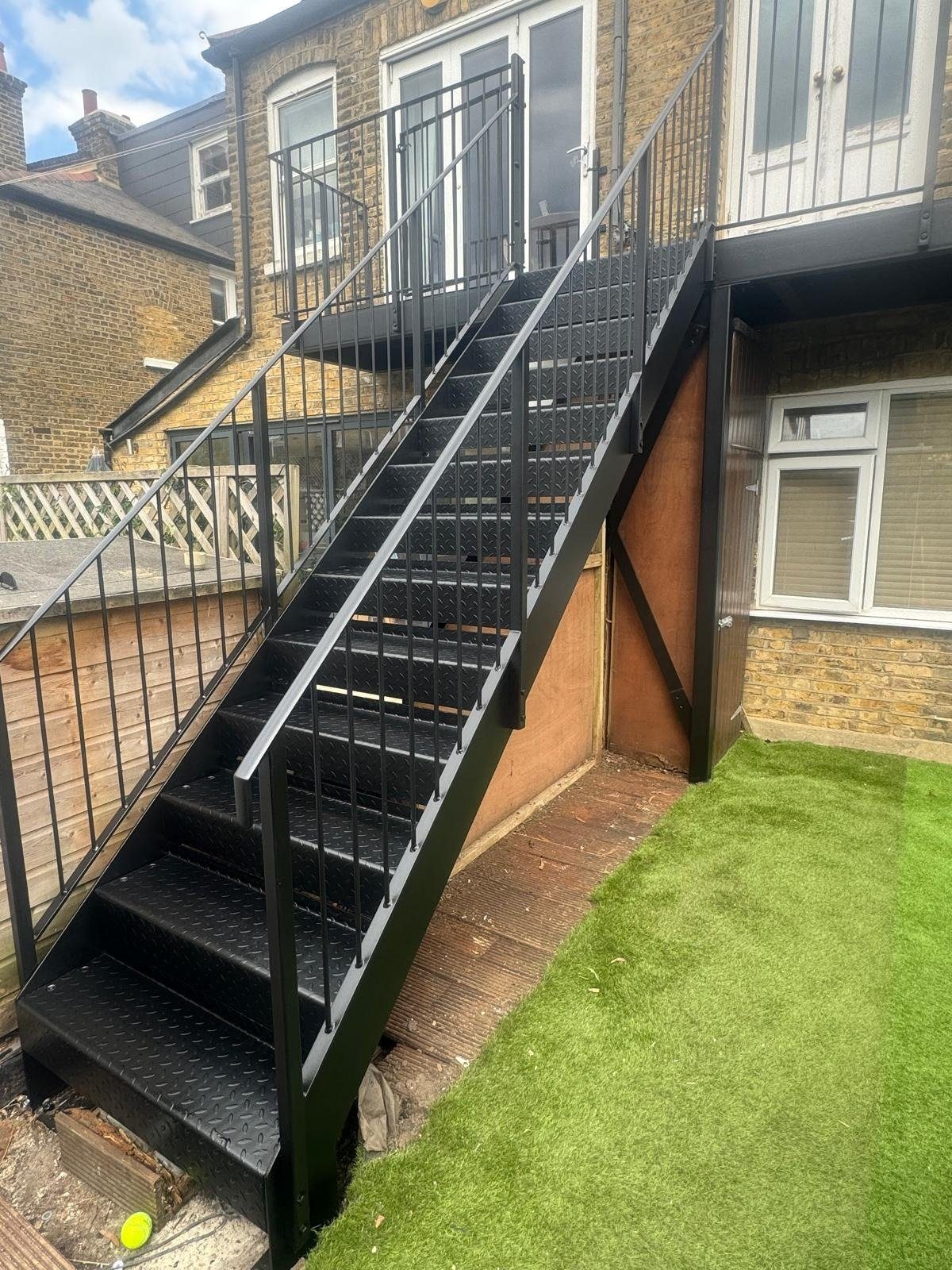Black steel external staircase with chequer plate treads and vertical bar balustrade rising from an artificial lawn to a first-floor balcony at the rear of a brick house