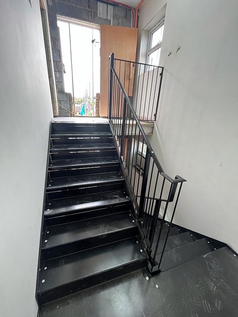 Black steel staircase with open perforated treads and a quarter landing, stainless steel handrail, in a bright showroom with white tiled wall, timber floor and full-height glazing