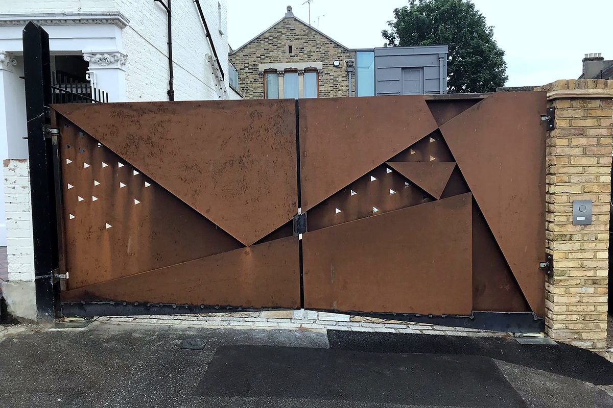 Corten-look steel double driveway gates with angled sheet panels and small triangle cut-outs, between a black steel post and a yellow-brick pier