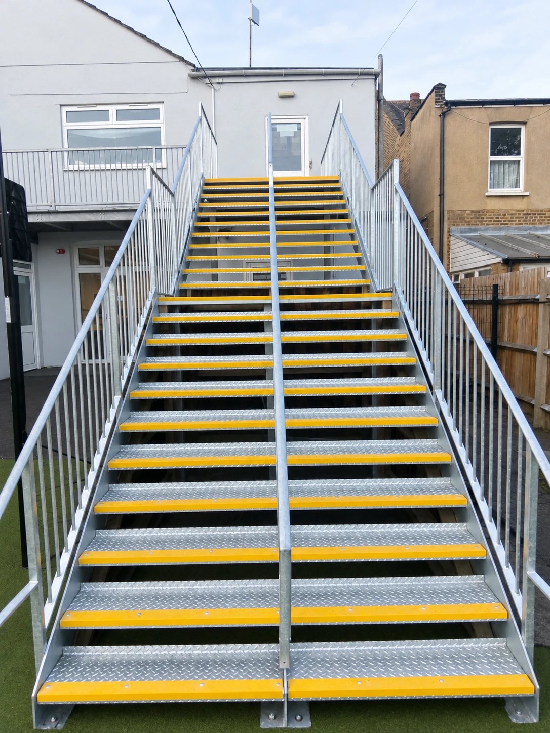 Wide hot-dip galvanised staircase with chequer-plate treads, yellow nosings and a centre handrail, rising to a first-floor door