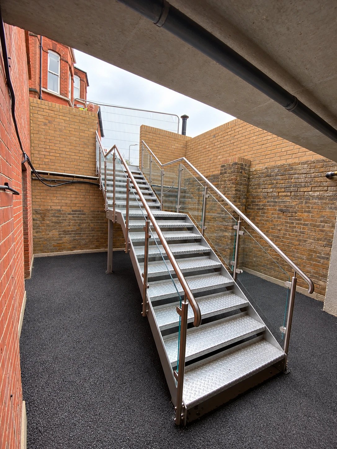 Galvanised steel staircase with chequer-plate treads, glass balustrade and stainless steel handrail rising out of a brick-walled courtyard