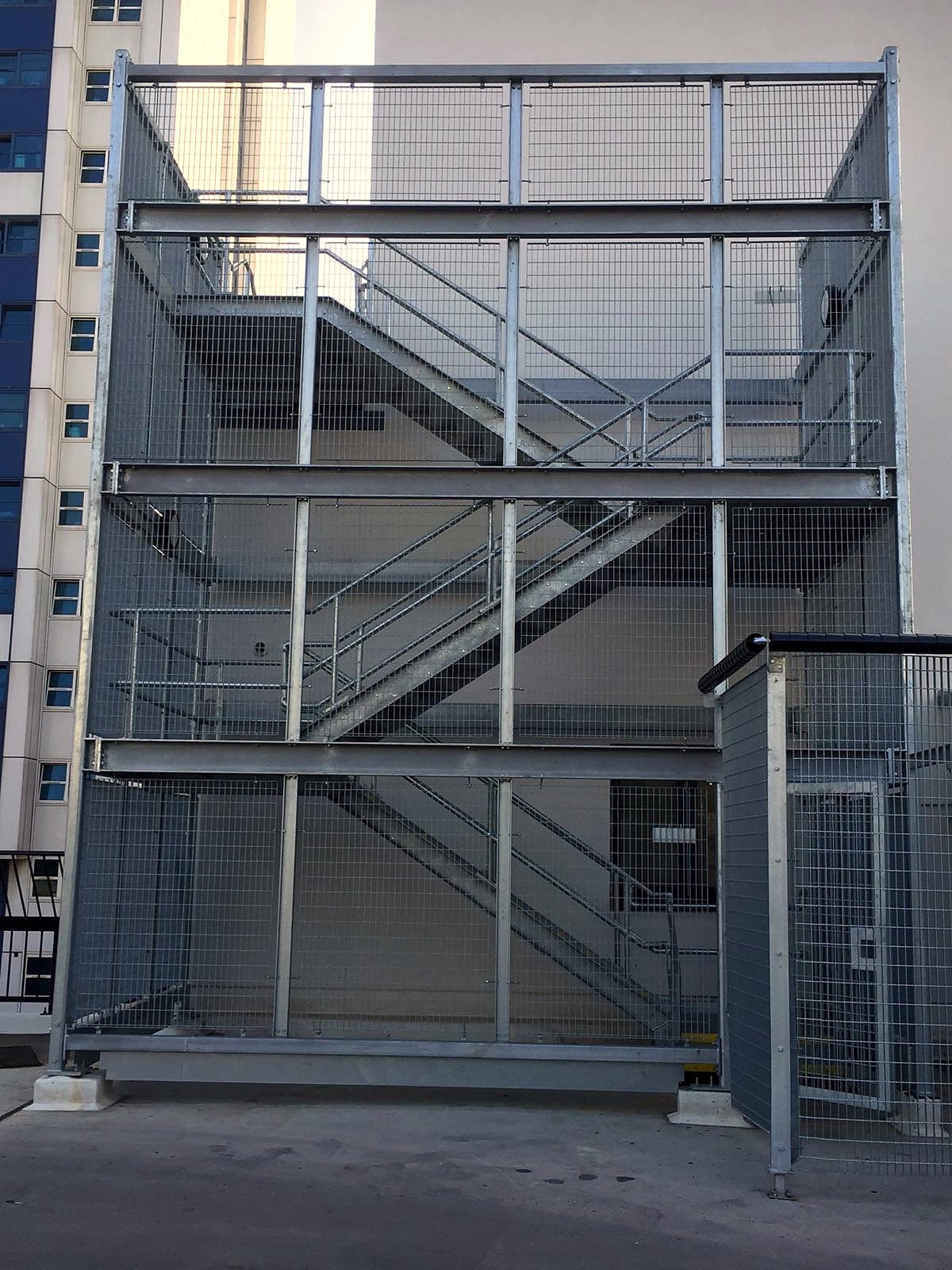 Galvanised steel stair tower with welded mesh infill panels beside a commercial building