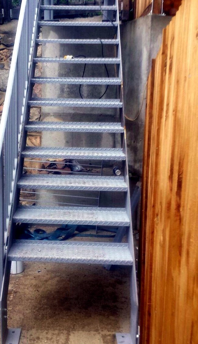 Galvanised steel staircase with bar railings rising between a stone wall and a timber fence