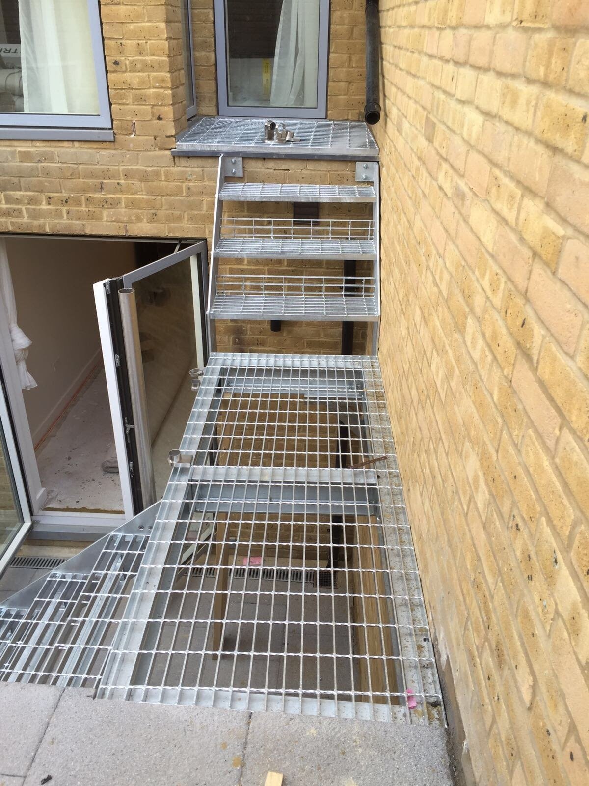 Galvanised steel walkway and short flight of steps with mesh balustrade linking doorways in a brick lightwell