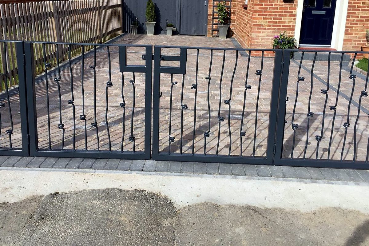 Grey steel double driveway gates with wavy vertical bars and small coiled knots, across a block-paved drive in front of a red-brick house