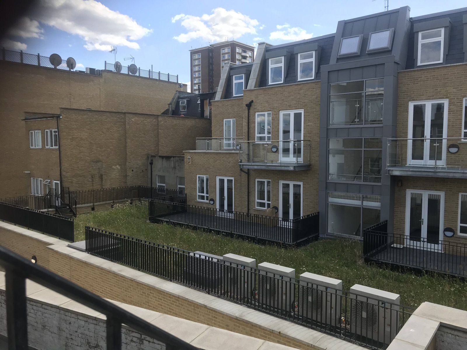 Rear of a brick apartment block with steel balconies and black boundary railings around a communal garden