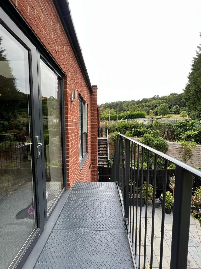 Grey steel balcony walkway with chequer plate deck and vertical bar railings along the rear of a brick extension